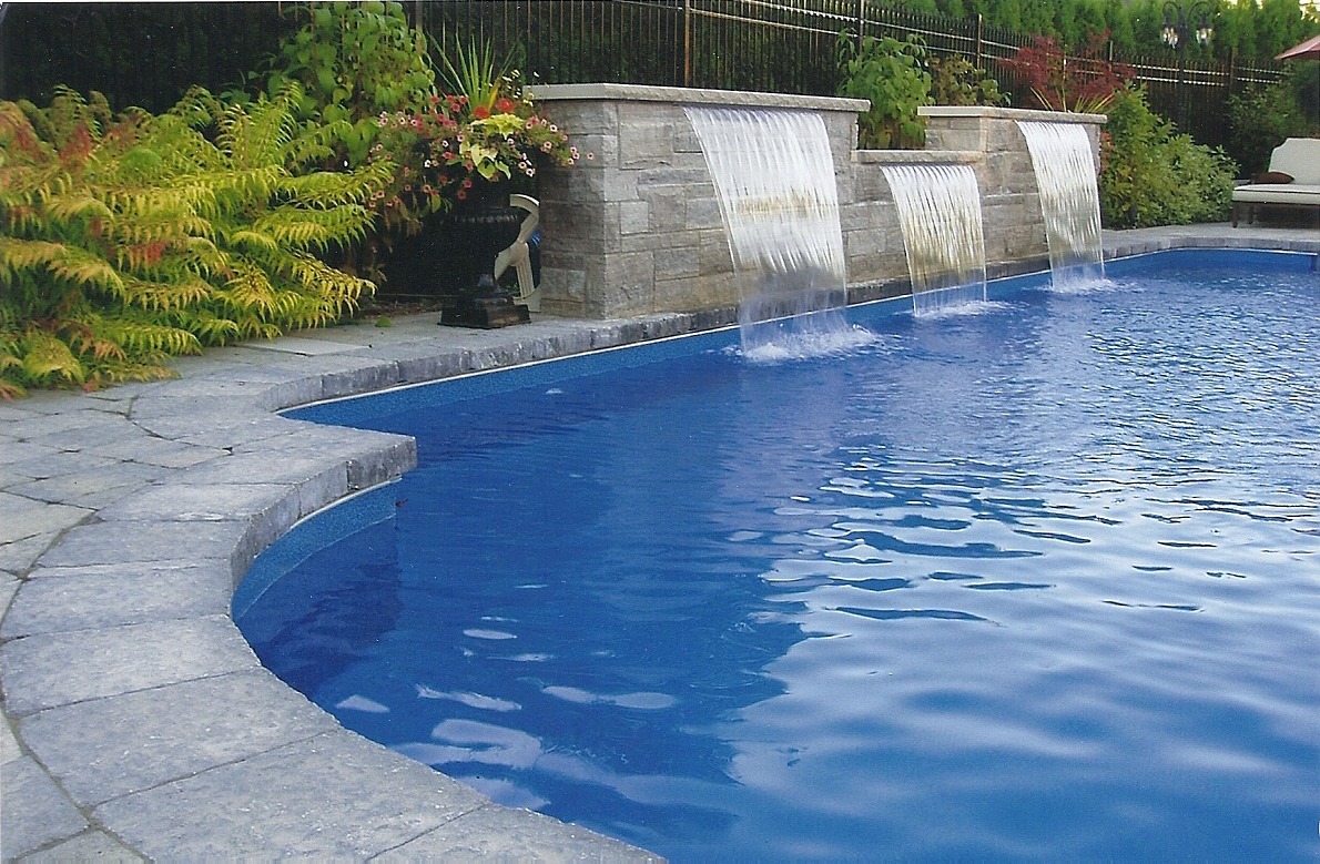 A serene swimming pool with cascading waterfalls, surrounded by lush green plants and stone tiles, creates a tranquil backyard oasis.